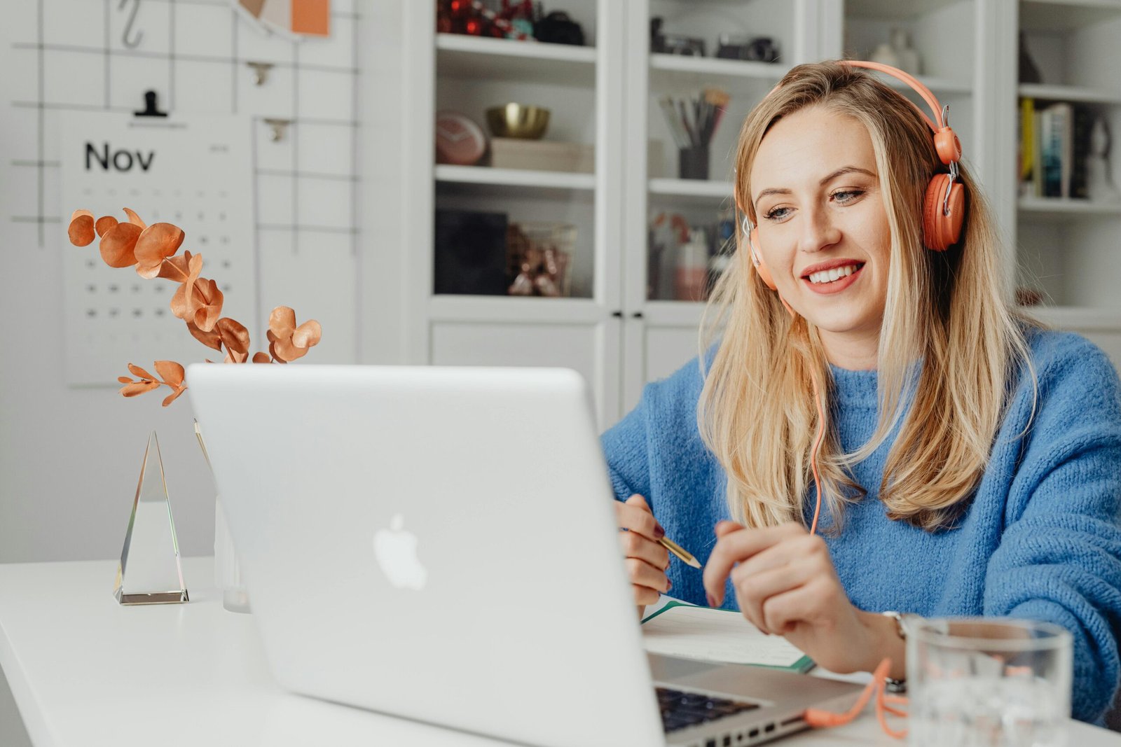 Blonde woman smiling while working remotely on a laptop, wearing headphones in a cozy home office.