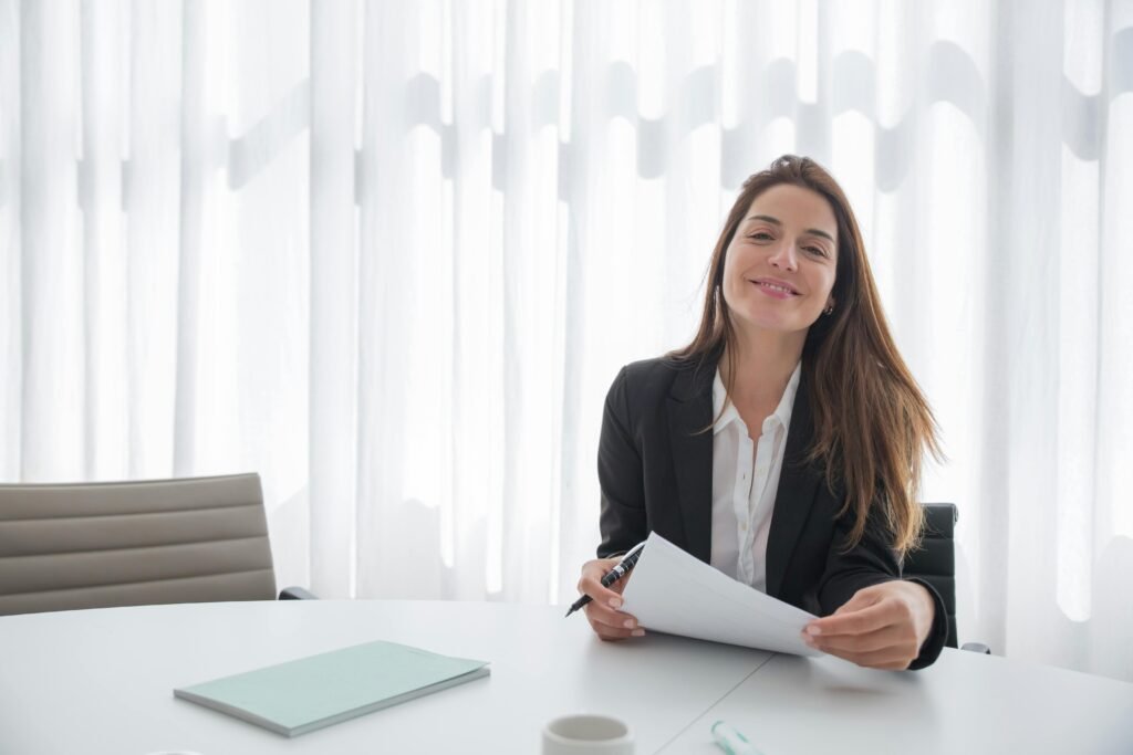 Smiling businesswoman in a blazer holding paper at an office table indoors.